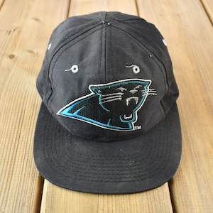 Op de afbeelding: Zwarte en blauwe baseballpet met een Carolina Panthers logo. Het logo is een zwarte panter met een blauwe omtrek.