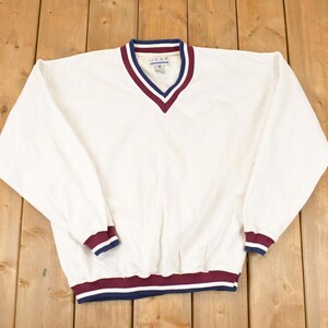 Puede incluir: Un jersey blanco con cuello en V y mangas largas. El cuello, los puños y el dobladillo presentan un diseño a rayas azul marino y granate. El jersey está hecho de un material ligero.