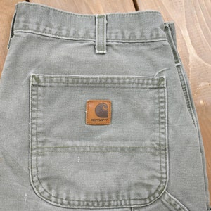 May include: A pair of green Carhartt work trousers with a large pocket on the back. The pocket has a brown Carhartt logo patch.