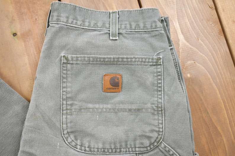 May include: A pair of green Carhartt work trousers with a large pocket on the back. The pocket has a brown Carhartt logo patch.