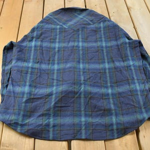 May include: A blue plaid shirt with long sleeves. The shirt has a classic plaid pattern in shades of blue, green, and brown. The shirt is laid flat on a wooden surface, showing the back of the garment.