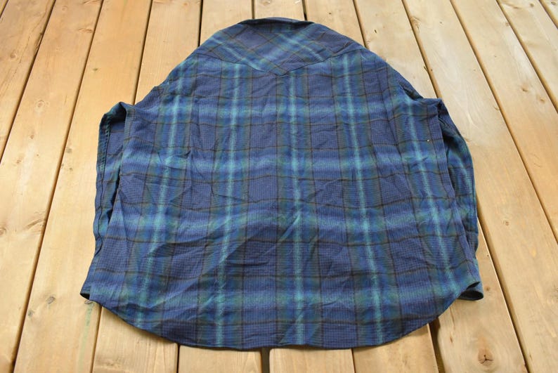 May include: A blue plaid shirt with long sleeves. The shirt has a classic plaid pattern in shades of blue, green, and brown. The shirt is laid flat on a wooden surface, showing the back of the garment.