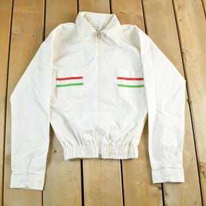 May include: White zip-up jacket with a collar and long sleeves. The jacket has two pockets, each adorned with a horizontal red and green stripe. The bottom hem is elasticized.