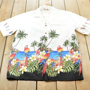 Könnte beinhalten: Weißes Hemd mit Knöpfen und tropischem Design. Das Hemd hat einen farbenfrohen Rand mit Papageien, Palmen, Blumen und einer blauen Ozeanszene. Die Ärmel haben kleine Fischmuster.