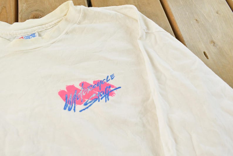 May include: Cream-colored long-sleeve t-shirt with a crew neck. The shirt features a graphic in pink and blue that reads "Motorcycle Staff". The shirt is made of a soft, lightweight material.