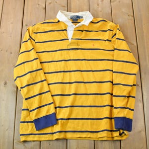 May include: A yellow and navy blue striped long-sleeved rugby shirt with a white collar and button placket. The shirt has a small embroidered logo on the left chest.