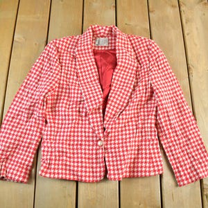 May include: A red and white houndstooth blazer with a single-button closure. The blazer has a notched collar and a red lining. The garment is laid flat on a wooden surface.