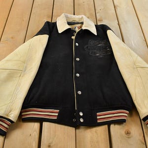 May include: A black varsity jacket featuring cream-coloured sleeves, a cream collar, and a front button closure. The jacket includes a black embroidered logo on the chest and ribbed cuffs and hem with red, white, and black striped detailing.