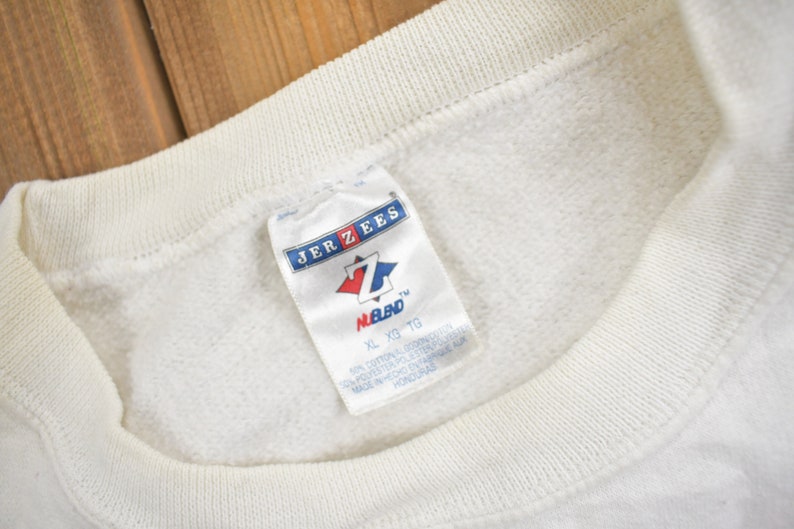 K&ouml;nnte beinhalten: Wei&szlig;er Jerzees-Sweatshirt mit einem Etikett mit der Aufschrift "XL XG TG" und "M.B.D."