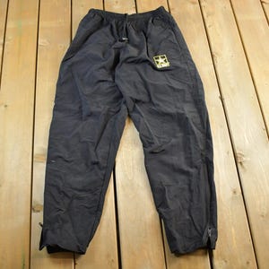 Puede incluir: Pantalones deportivos negros de nailon con cintura elástica y cremalleras en los tobillos. Un parche con el logotipo del ejército dorado y negro está en el muslo izquierdo. Los pantalones están sobre una superficie de madera.