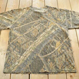 Puede incluir: Una camiseta de camuflaje de manga corta con cuello redondo. La camiseta presenta un patr&oacute;n de color marr&oacute;n, beige y verde, que se asemeja a una escena de bosque o arboleda. La camiseta est&aacute; hecha de un material suave.