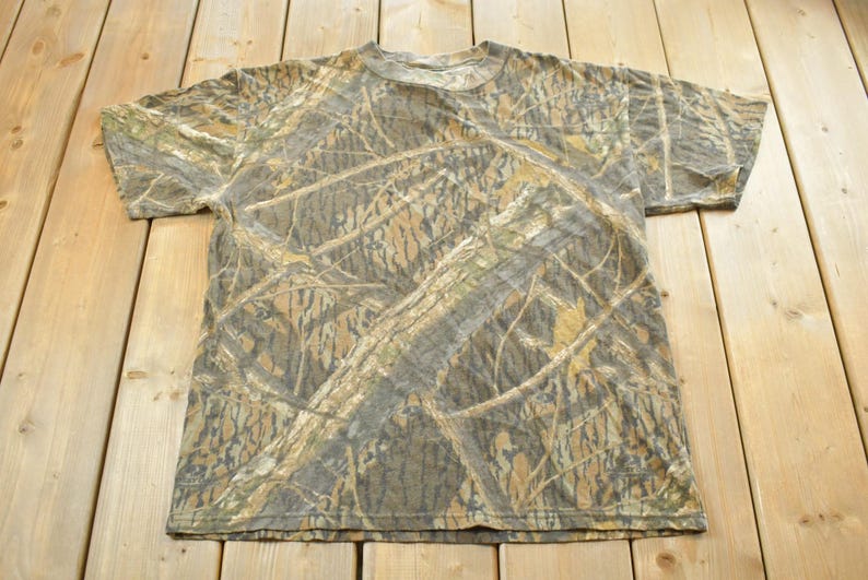 Puede incluir: Una camiseta de camuflaje de manga corta con cuello redondo. La camiseta presenta un patr&oacute;n de color marr&oacute;n, beige y verde, que se asemeja a una escena de bosque o arboleda. La camiseta est&aacute; hecha de un material suave.