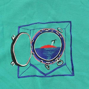 May include: Teal-colored fabric with an embroidered design of a ship's porthole. The porthole is open, revealing a red island, blue water, and a small palm tree. The porthole is framed in blue, with the open door on the left.