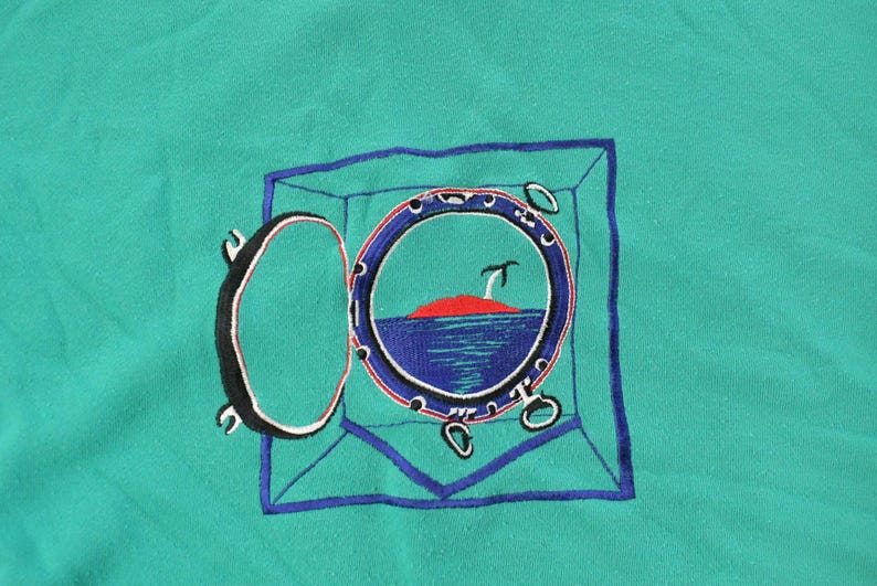 May include: Teal-colored fabric with an embroidered design of a ship's porthole. The porthole is open, revealing a red island, blue water, and a small palm tree. The porthole is framed in blue, with the open door on the left.