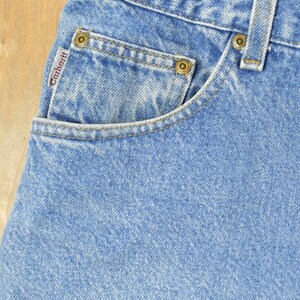 Puede incluir: Primer plano de vaqueros de mezclilla azul claro con un bolsillo y la etiqueta de la marca Carhartt. Los vaqueros tienen remaches de color lat&oacute;n y un dise&ntilde;o cl&aacute;sico de cinco bolsillos. La tela tiene un aspecto texturizado.