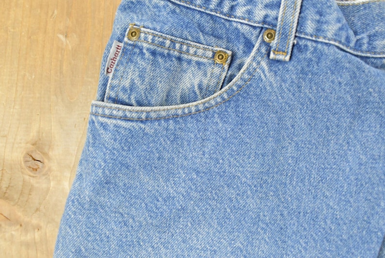 Puede incluir: Primer plano de vaqueros de mezclilla azul claro con un bolsillo y la etiqueta de la marca Carhartt. Los vaqueros tienen remaches de color lat&oacute;n y un dise&ntilde;o cl&aacute;sico de cinco bolsillos. La tela tiene un aspecto texturizado.