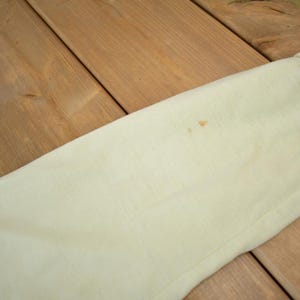 May include: A close-up of a light cream colored fabric with a small brown stain. The fabric is smooth and appears to be made of cotton.