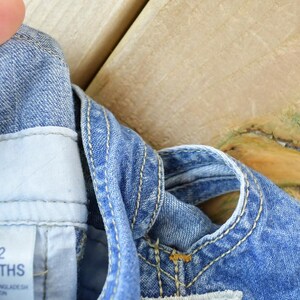 May include: Close-up of a denim overall garment for a baby. The blue denim has white stitching and a tag that reads "12 MONTHS". The garment is made of cotton.