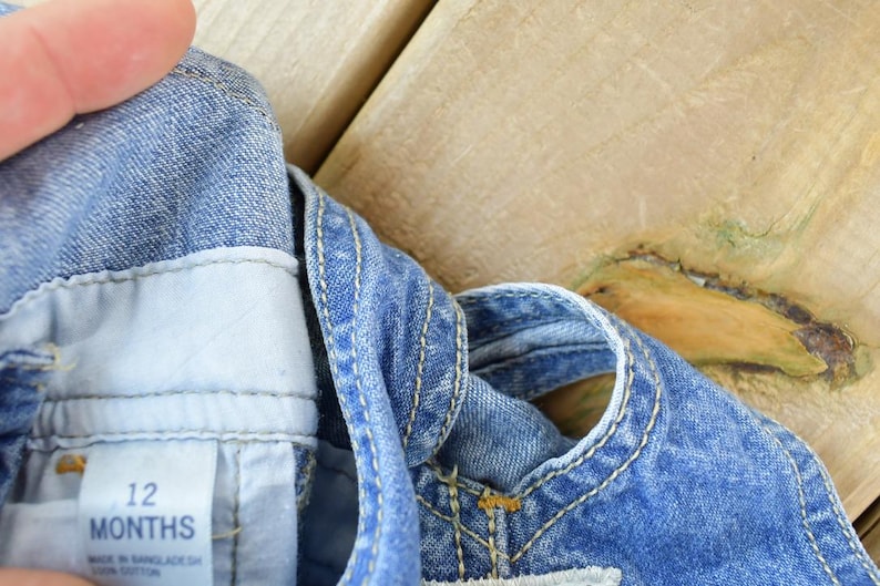 May include: Close-up of a denim overall garment for a baby. The blue denim has white stitching and a tag that reads "12 MONTHS". The garment is made of cotton.