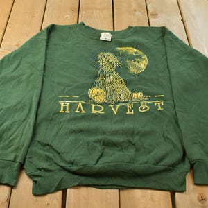 May include: A dark green crewneck sweatshirt featuring a yellow graphic print of a harvest scene with a haystack, pumpkins, and a full moon. The word HARVEST is printed in yellow capital letters below the central illustration on the front.