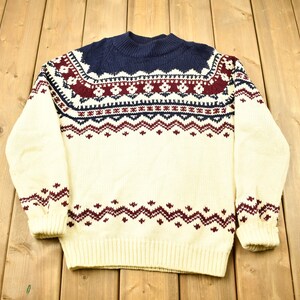 Könnte beinhalten: Ein cremefarbener Pullover mit einem roten, blauen und weißen geometrischen Muster. Der Pullover hat einen Rollkragen und lange Ärmel.