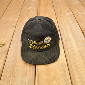 Könnte beinhalten: Schwarze Cord-Baseballkappe mit dem Pittsburgh Steelers-Logo, das in Gold, Weiß und Gelb gestickt ist. Die Kappe hat einen gebogenen Schirm und einen Knopf oben. Ein klassisches Sportaccessoire.