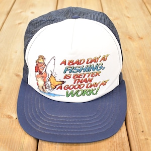 Puede incluir: Gorra de camionero azul marino y blanca con el texto "¡Un mal día de pesca es mejor que un buen día de trabajo!" y un dibujo de pescador. La gorra tiene una parte trasera de malla.