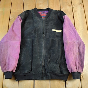 May include: Black bomber jacket with purple sleeves. The jacket has a zipper closure and a ribbed collar and cuffs. The word "Vikings" is printed on the chest. The jacket is made of a suede-like material.