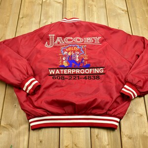 Può includere: Giubbotto bomber rosso in satin con bordature a righe bianche e rosse. Sul retro del giubbotto è presente un logo ricamato che dice "Jacoby Waterproofing" con un personaggio dei cartoni animati che tiene un secchio e un telefono. Il telefono dice "Aiuto!" su di esso.