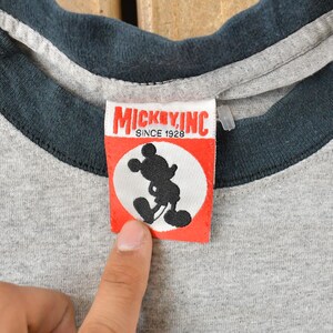 May include: Close-up of a gray ringer t-shirt with a dark blue collar and sleeve trim. A red and white tag features a black silhouette of Mickey Mouse and the text "MICKEY, INC. SINCE 1928".