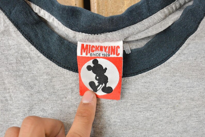 May include: Close-up of a gray ringer t-shirt with a dark blue collar and sleeve trim. A red and white tag features a black silhouette of Mickey Mouse and the text "MICKEY, INC. SINCE 1928".