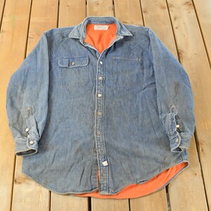 Könnte beinhalten: Ein blaues Jeanshemd mit orangefarbenem Futter. Das langärmlige Hemd hat zwei Brusttaschen und eine Knopfleiste. Kragen und Manschetten sind ebenfalls geknöpft. Das Hemd liegt flach auf einer Holzoberfläche.