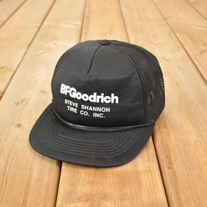 Könnte beinhalten: Schwarze Trucker-Kappe mit dem BFGoodrich-Logo und dem weißen Text "STEVE SHANNON TIRE CO. INC.". Die Kappe hat einen Mesh-Rücken und einen gebogenen Schirm. Auf einer Holzoberfläche platziert.