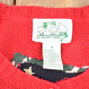 K&ouml;nnte beinhalten: Roter Strickpullover mit gr&uuml;nem, wei&szlig;em und schwarzem Besatz am Ausschnitt. Der Pullover hat eine goldene Stern-Applikation. Das am Pullover befestigte Etikett lautet "The Quacker Factory" und listet die Stoffzusammensetzung als 55% Ramie, 45% Baumwolle und 70% Lammwolle auf.