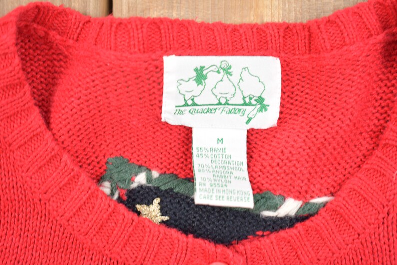 K&ouml;nnte beinhalten: Roter Strickpullover mit gr&uuml;nem, wei&szlig;em und schwarzem Besatz am Ausschnitt. Der Pullover hat eine goldene Stern-Applikation. Das am Pullover befestigte Etikett lautet "The Quacker Factory" und listet die Stoffzusammensetzung als 55% Ramie, 45% Baumwolle und 70% Lammwolle auf.