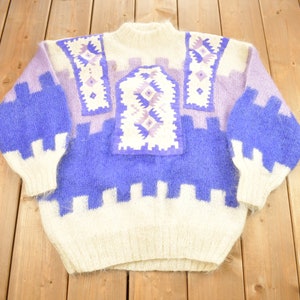 May include: A white and purple fuzzy knit sweater with a geometric pattern. The sweater has a high neck and long sleeves.