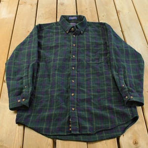 Peut inclure: Chemise à manches longues à carreaux verts, marine et jaunes. Elle présente un col boutonné, une poche poitrine et des poignets boutonnés. Le tissu est doux, peut-être en flanelle. La chemise est posée à plat.