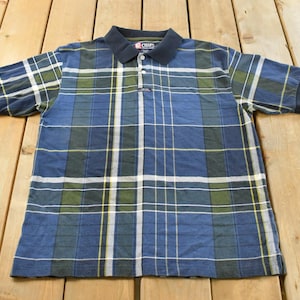 May include: A short-sleeve polo shirt featuring a blue, green, and white plaid pattern. The shirt has a solid navy blue collar and sleeve cuffs, a three-button placket, and a small Chaps brand label located on the chest area of the garment.