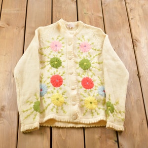 May include: A cream-colored cardigan sweater with embroidered flowers in shades of pink, yellow, green, and blue. The cardigan has a button-up front.