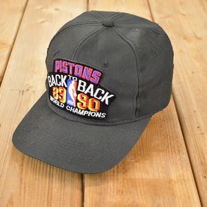 Può includere: Un cappellino da baseball nero con visiera curva. Il cappellino presenta una toppa con la scritta "Pistons Back to Back 89 90 World Champions" in rosso, bianco e blu. Il cappellino è su uno sfondo di legno.