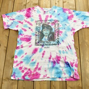 May include: A white tie-dye t-shirt with a blue and pink design. The shirt features a graphic of a woman with a blue beret and glasses. The text on the shirt reads "But lation tow sum sled chenic se po CAMC THUL Das med her we dane LOWONE".