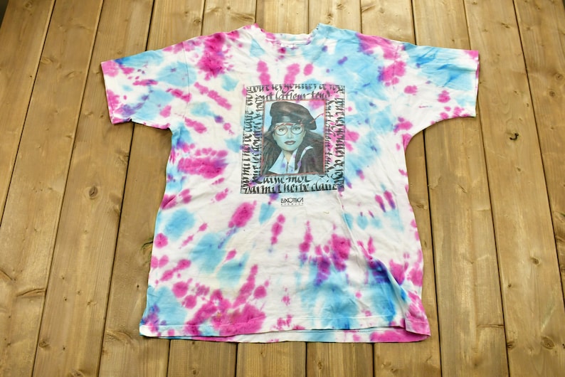 May include: A white tie-dye t-shirt with a blue and pink design. The shirt features a graphic of a woman with a blue beret and glasses. The text on the shirt reads "But lation tow sum sled chenic se po CAMC THUL Das med her we dane LOWONE".