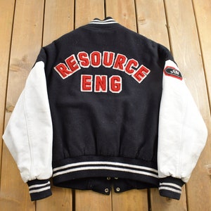 Puede incluir: Chaqueta de béisbol negra y blanca con letras rojas que dicen "RESOURCE ENG" en la espalda. Las mangas son de cuero blanco y la chaqueta tiene un ribete de rayas blancas y negras alrededor de la parte inferior y los puños.