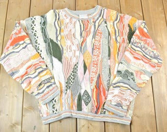 May include: A colorful, patterned sweater with a crew neck. The sweater features a variety of textures and designs in shades of orange, yellow, green, and gray. The sleeves and hem have a light gray trim.