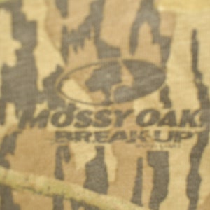 May include: Camouflage pattern fabric with the "Mossy Oak Break-Up" logo. The design features shades of brown, tan, and green, creating a naturalistic, outdoor-themed aesthetic. The logo is in a dark oval.
