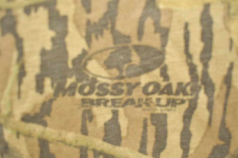 May include: Camouflage pattern fabric with the "Mossy Oak Break-Up" logo. The design features shades of brown, tan, and green, creating a naturalistic, outdoor-themed aesthetic. The logo is in a dark oval.