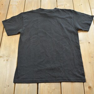 May include: A black t-shirt, seen from the back, rests on a light brown wooden surface. The shirt features short sleeves and a crew neck. The fabric has a slightly worn appearance.
