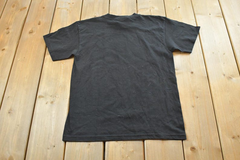 May include: A black t-shirt, seen from the back, rests on a light brown wooden surface. The shirt features short sleeves and a crew neck. The fabric has a slightly worn appearance.
