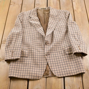 Può includere: Un blazer a quadri marroni e beige con chiusura a un bottone. Il blazer ha un revers a punta e due tasche anteriori.