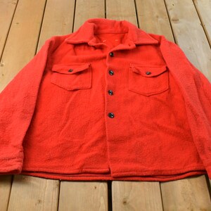 May include: A bright red long-sleeve shirt made of a thick, textured wool-like fabric. This button-down garment features a pointed collar, two chest pockets with buttoned flaps, and dark buttons running down the centre front placket.